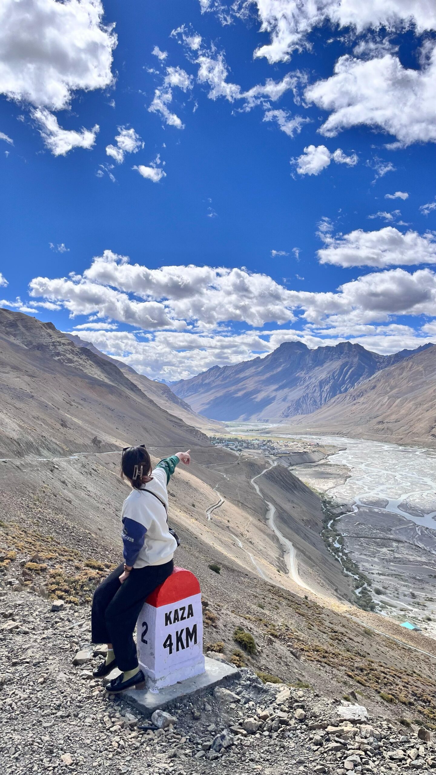I am posing beside a milestone marked Kaza 4km, under a blue sky with scattered clouds, witnessing the scenic beauty on the way to the key monastery in Spiti